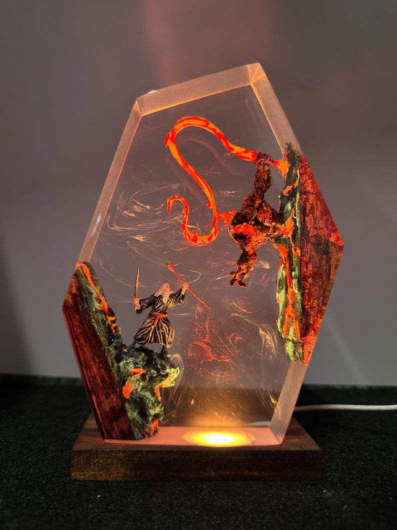 Puede incluir: L&aacute;mpara decorativa con una escena de fantas&iacute;a encapsulada en resina transparente. La escena muestra dos figuras en combate, con un elemento naranja fuego. La l&aacute;mpara tiene una base de madera y emite un brillo c&aacute;lido. La resina tiene forma geom&eacute;trica.