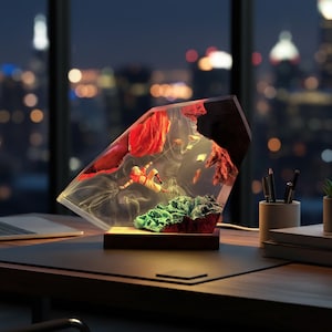 May include: A decorative lamp with a clear, geometric resin body, showcasing red and green elements within. The lamp sits on a wooden base, illuminated from within, and is placed on a desk with a laptop and city lights in the background.