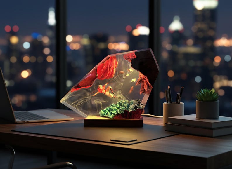 May include: A decorative lamp with a clear, geometric resin body, showcasing red and green elements within. The lamp sits on a wooden base, illuminated from within, and is placed on a desk with a laptop and city lights in the background.