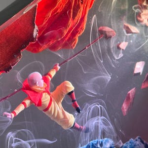 May include: A miniature figure in a yellow and red outfit, holding a staff, appears to be floating in a smoky, abstract environment. Red and blue elements frame the scene, with floating rock-like shapes.