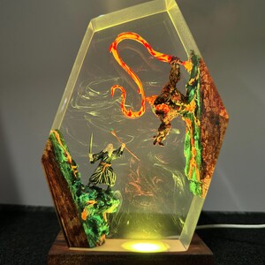Puede incluir: L&aacute;mpara hexagonal de resina que representa una escena de fantas&iacute;a con un mago, una criatura y un elemento de fuego. La l&aacute;mpara tiene una base de madera y un brillo amarillo c&aacute;lido. Las figuras est&aacute;n sobre un fondo de colores arremolinados.