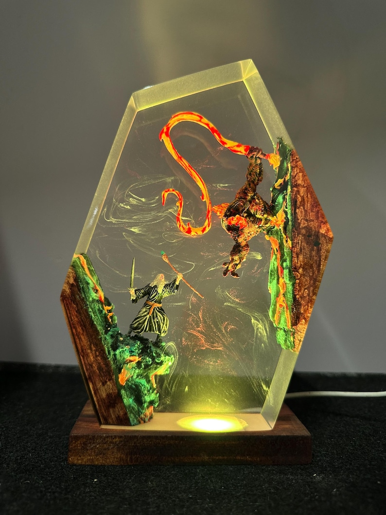 Puede incluir: L&aacute;mpara hexagonal de resina que representa una escena de fantas&iacute;a con un mago, una criatura y un elemento de fuego. La l&aacute;mpara tiene una base de madera y un brillo amarillo c&aacute;lido. Las figuras est&aacute;n sobre un fondo de colores arremolinados.