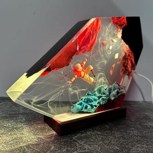 May include: A unique resin lamp featuring a miniature figure in an orange suit climbing a red rock formation. The lamp has a wooden base and a clear, angled body with a black top. The scene includes blue and red elements.