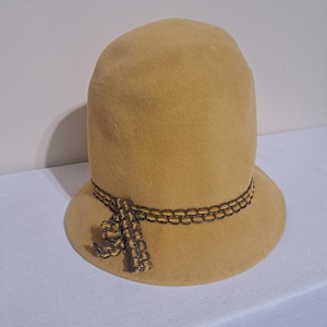 Puede incluir: Un sombrero cloche color beige con una copa redondeada y un ala pequeña inclinada hacia abajo. El sombrero está adornado con una banda decorativa de adorno en forma de cadena marrón oscuro y una borla a juego. El sombrero es de fieltro suave.