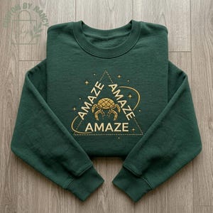 May include: A dark green sweatshirt with long sleeves. The front displays a gold embroidered design with the word "AMAZE" repeated around a geometric shape and a crab-like figure. The sweatshirt is folded on a wooden surface.