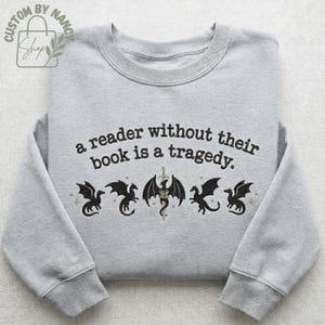 Może przedstawiać: Jasnoszara bluza z haftowanym napisem "a reader without their book is a tragedy". Poniżej tekstu znajduje się pięć czarnych wzor&oacute;w smok&oacute;w ze złotymi akcentami. Bluza ma okrągły dekolt i długie rękawy.