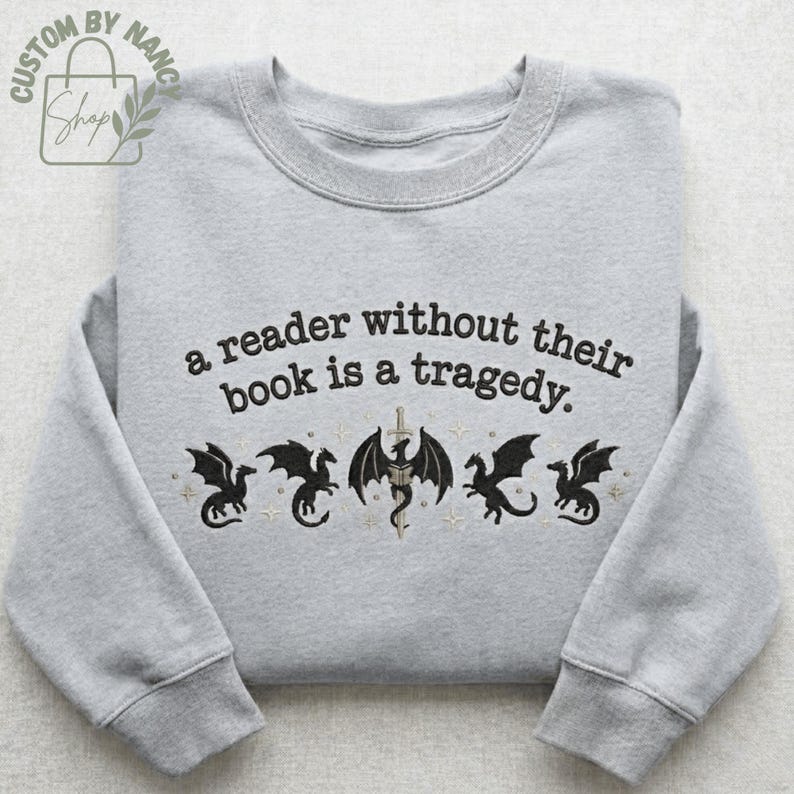 Może przedstawiać: Jasnoszara bluza z haftowanym napisem "a reader without their book is a tragedy". Poniżej tekstu znajduje się pięć czarnych wzor&oacute;w smok&oacute;w ze złotymi akcentami. Bluza ma okrągły dekolt i długie rękawy.