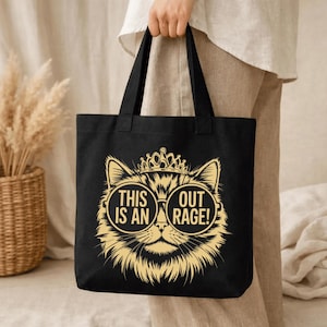 May include: Black tote bag featuring a gold illustration of a cat wearing sunglasses and a crown. The text on the bag reads "THIS IS AN OUT RAGE!". The bag has two black handles.