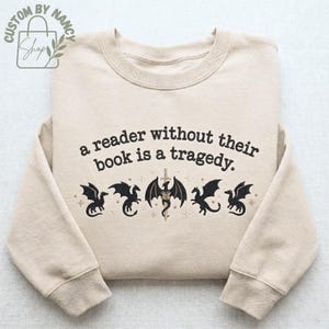 Może przedstawiać: Beżowa bluza z napisem "a reader without their book is a tragedy". Poniżej tekstu znajduje się pięć czarnych wzor&oacute;w smok&oacute;w. Bluza ma długie rękawy i okrągły dekolt.