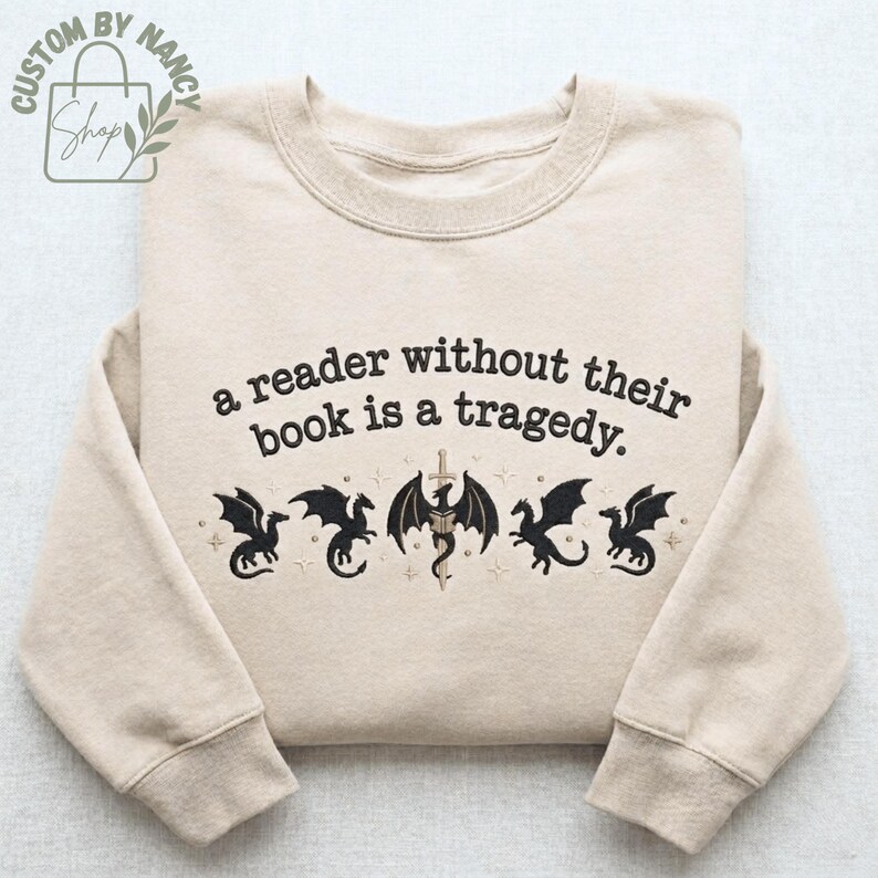 Może przedstawiać: Beżowa bluza z napisem "a reader without their book is a tragedy". Poniżej tekstu znajduje się pięć czarnych wzor&oacute;w smok&oacute;w. Bluza ma długie rękawy i okrągły dekolt.
