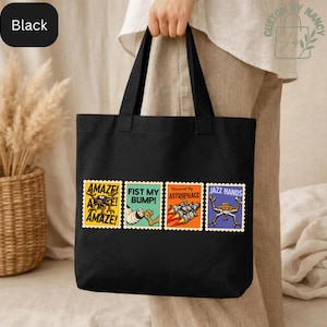 May include: Black tote bag featuring four colorful postage stamp designs with the phrases "AMAZE!", "FIST MY BUMP!", "Powered By Astrophage", and "JAZZ HANDS". The bag is held by a person.