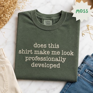 May include: A moss green crew neck t-shirt with the embroidered text "does this shirt make me look professionally developed" in a light cream color. The shirt features a Comfort Colors brand label on the inside of the collar.