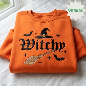 May include: An orange crewneck sweatshirt featuring black embroidered text that reads Witchy. The design includes a black witch hat with a gold buckle, a tan broomstick, two black bats, and several small four-pointed star accents.