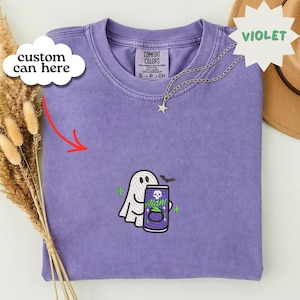May include: A violet short-sleeve t-shirt featuring an embroidered graphic of a white ghost holding a purple soda can with a skull and cauldron design. The shirt has a crew neckline and a Comfort Colors label at the inner collar. The text custom can here is visible in a white cloud graphic, and the word VIOLET is printed in a white starburst shape.