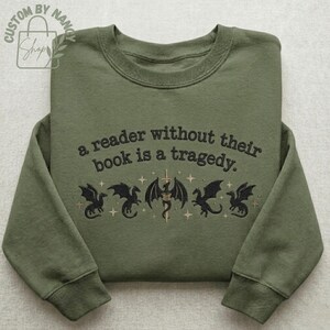Może przedstawiać: Zielona bluza z haftowanym napisem: "a reader without their book is a tragedy". Poniżej znajduje się pięć czarnych smok&oacute;w z mieczem i gwiazdami. Bluza ma okrągły dekolt i długie rękawy.
