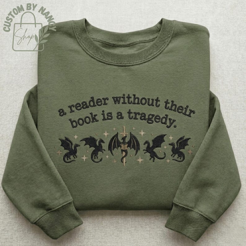 Może przedstawiać: Zielona bluza z haftowanym napisem: "a reader without their book is a tragedy". Poniżej znajduje się pięć czarnych smok&oacute;w z mieczem i gwiazdami. Bluza ma okrągły dekolt i długie rękawy.