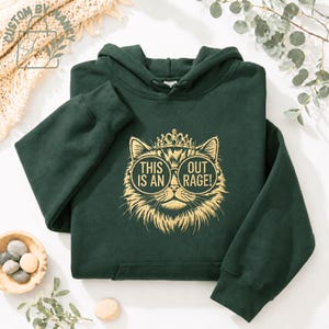May include: A dark green hoodie featuring a cat design with sunglasses and a crown. The cat graphic includes the text "THIS IS AN OUT RAGE!". The hoodie has a front pocket and a hood.