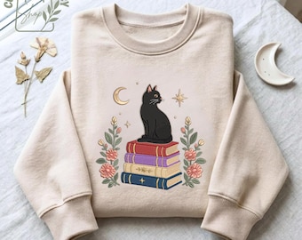Könnte beinhalten: Ein beigefarbenes Sweatshirt mit Rundhalsausschnitt und einem Grafikdruck einer schwarzen Katze, die auf einem Stapel aus vier bunten Büchern sitzt. Das Design umfasst florale Akzente auf beiden Seiten, eine Mondsichel und einen Stern.