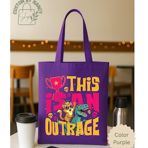 May include: Purple tote bag with the text "THIS IS AN OUTRAGE" in yellow and pink. The design features a cat wearing sunglasses and a dinosaur, with a trophy and skulls. The bag has purple handles and is on a white surface.