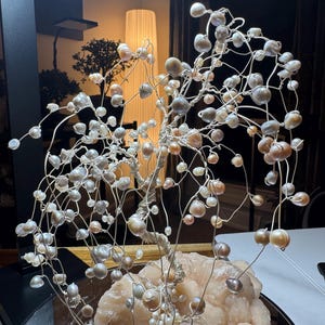 May include: A handcrafted wire tree sculpture adorned with numerous pearls in shades of white, gray, and peach. The tree is mounted on a light pink, textured stone base, creating a unique decorative piece.