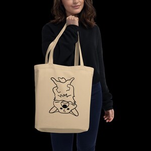 May include: A beige tote bag with a black outline drawing of a dog, upside down. The dog's face and paws are visible. The bag is held by a person wearing a black sweater. The bag is made of a natural material.