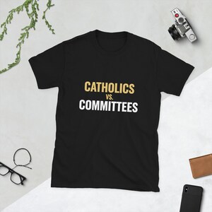 Op de afbeelding: Zwart T-shirt met de tekst "CATHOLICS vs. COMMITTEES" in gouden en witte letters. Het shirt heeft een ronde hals en korte mouwen.