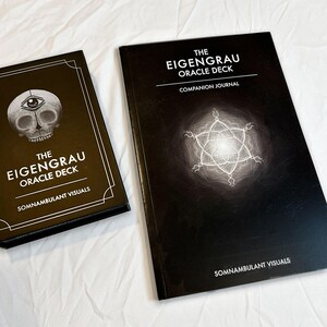 May include: Two black oracle deck products on a white surface. The deck on the left features a skull with an eye, and the text "THE EIGENGRAU ORACLE DECK" and "SOMNAMBULANT VISUALS". The deck on the right is a companion journal with a glowing design and the same text.