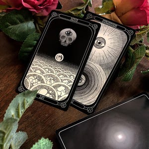 May include: A collection of tarot cards with black and white illustrations. One card features a skull with an eye, labeled "DEATH." Another card shows an eye surrounded by rays, labeled "IMAGINATION." The cards are set on a wooden surface with roses and greenery.