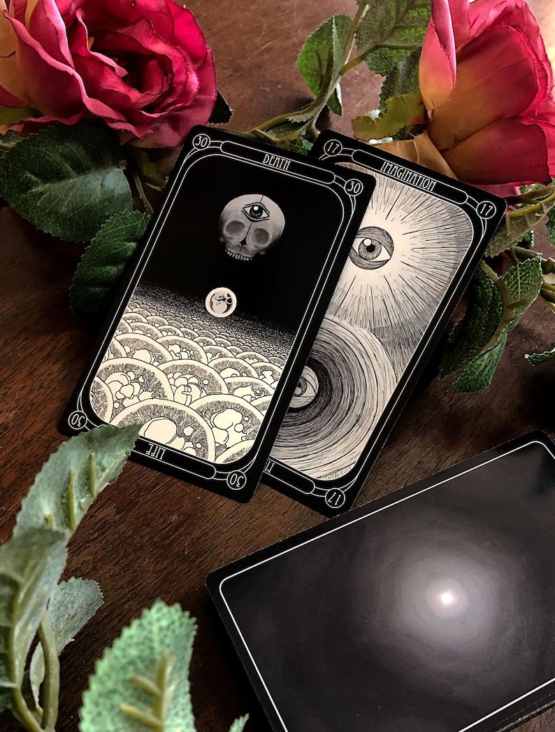 May include: A collection of tarot cards with black and white illustrations. One card features a skull with an eye, labeled "DEATH." Another card shows an eye surrounded by rays, labeled "IMAGINATION." The cards are set on a wooden surface with roses and greenery.