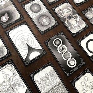 May include: A collection of black and white tarot cards with intricate illustrations. Each card features unique artwork, including geometric patterns, surreal figures, and symbolic imagery. The cards are arranged on a dark wooden surface.