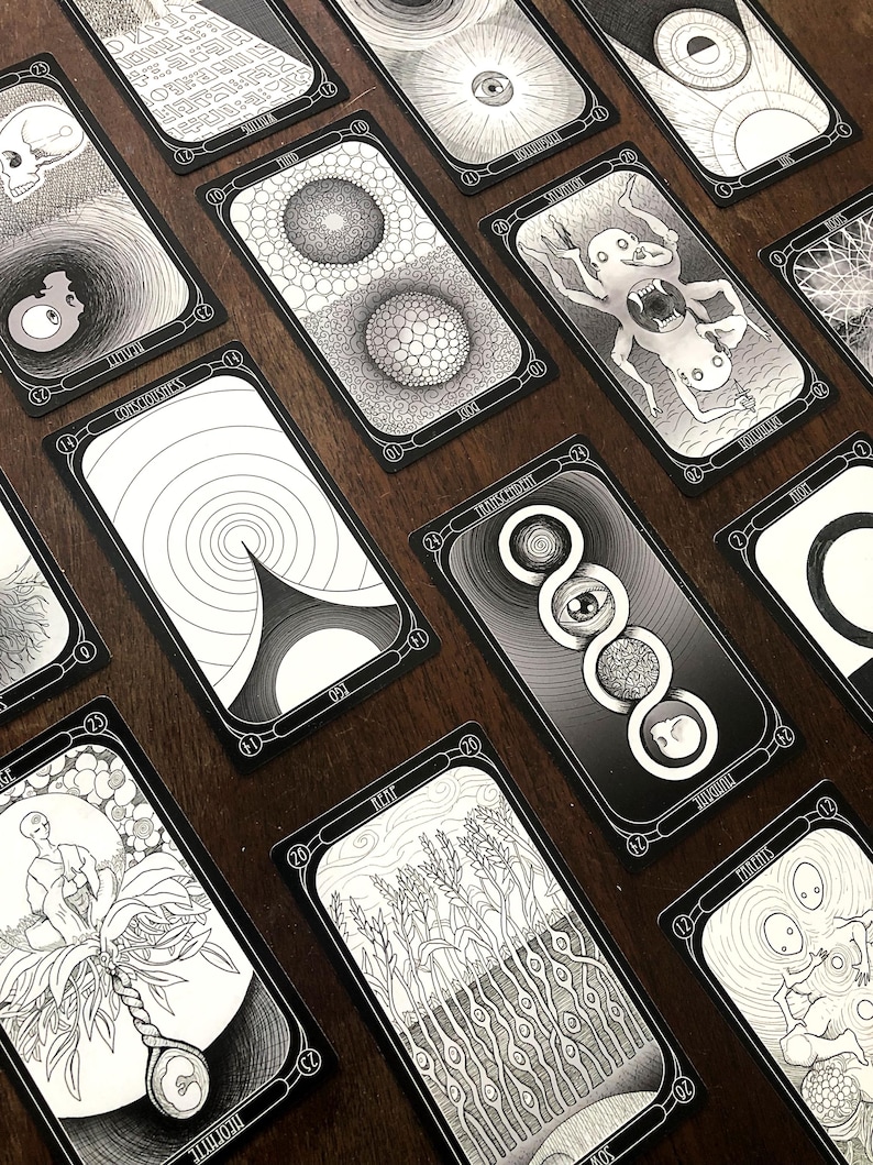 May include: A collection of black and white tarot cards with intricate illustrations. Each card features unique artwork, including geometric patterns, surreal figures, and symbolic imagery. The cards are arranged on a dark wooden surface.
