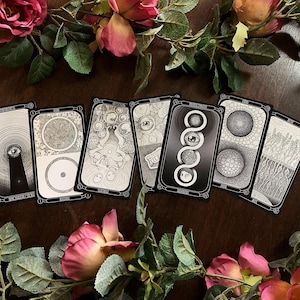 May include: A collection of tarot cards with black and white illustrations, arranged on a dark wooden surface. The cards feature various symbolic designs, including eyes, circles, and abstract patterns. Pink and red roses and green leaves frame the cards.