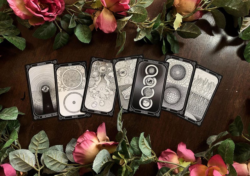 May include: A collection of tarot cards with black and white illustrations, arranged on a dark wooden surface. The cards feature various symbolic designs, including eyes, circles, and abstract patterns. Pink and red roses and green leaves frame the cards.