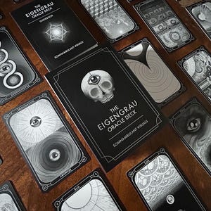 May include: The Eigenrau Oracle Deck, a divination tool, is shown with its guidebook and cards. The cards and guidebook are black with white illustrations and text. The deck features imagery of eyes, skulls, and geometric patterns.