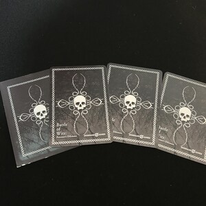 May include: Four playing cards with a dark background. Each card features a skull design with a cross-like pattern and the text "Battle of Wits: Poisoner's Dilemma" and "IRONMARK GAMES". The cards are black and white.