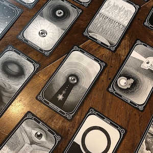 May include: A collection of black and white tarot cards with intricate illustrations and text. The cards are arranged on a wooden surface, showcasing various designs, including eyes, geometric patterns, and symbolic imagery. The cards are labeled with words like "Memory", "Awareness", and "Reading".