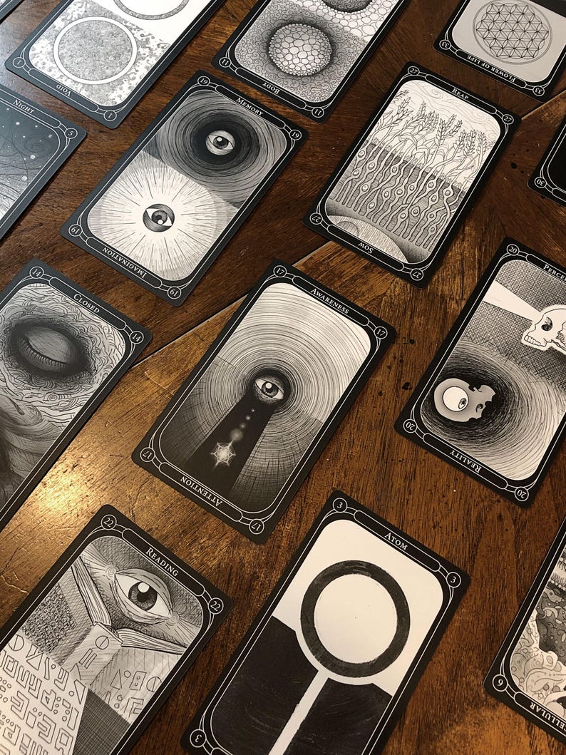 May include: A collection of black and white tarot cards with intricate illustrations and text. The cards are arranged on a wooden surface, showcasing various designs, including eyes, geometric patterns, and symbolic imagery. The cards are labeled with words like "Memory", "Awareness", and "Reading".
