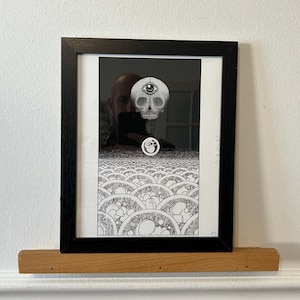 May include: A black framed print featuring a monochromatic illustration of a skull with a single eye on its forehead, positioned above a field of repeating circular patterns. The text "I to death" is written vertically on the right side.