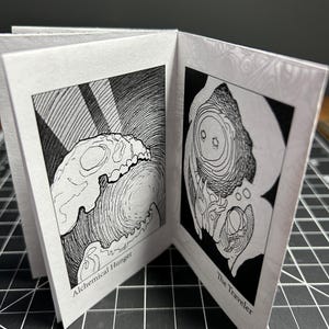 Puede incluir: Un pequeño libro abierto con dos ilustraciones en blanco y negro. La página izquierda presenta una imagen de calavera con el texto "Alchemical Hunger". La página derecha muestra una figura con el texto "The Traveler". El libro está sobre una superficie oscura.