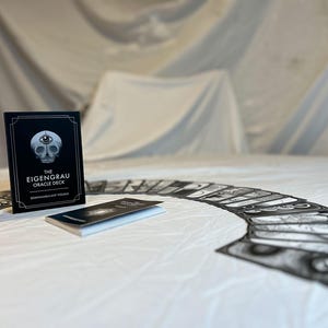 May include: The Eigenrau Oracle Deck, a divination tool, is displayed with its accompanying booklet and a spread of cards. The deck's box is black with white text and a skull illustration. The cards are arranged on a white surface.