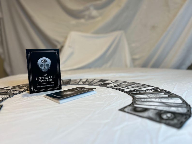 May include: The Eigenrau Oracle Deck, a divination tool, is displayed with its accompanying booklet and a spread of cards. The deck's box is black with white text and a skull illustration. The cards are arranged on a white surface.