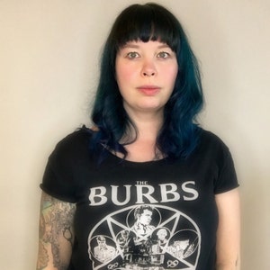 May include: A black short-sleeved t-shirt featuring a white graphic print of the movie title The Burbs with a central pentagram design containing various character illustrations and a house, set against a plain light-colored background.