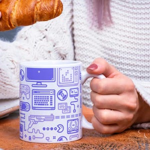 Puede incluir: Taza de cerámica blanca con un diseño de videojuego morado. La taza presenta ilustraciones de un monitor de computadora, teclado, controladores de juegos y otros iconos relacionados con los juegos. Un cruasán se sostiene en el fondo.