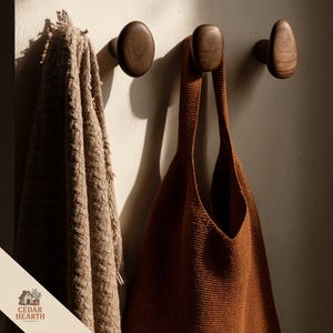 Może przedstawiać: Trzy ciemne, drewniane wieszaki ścienne są zamontowane na jasnej ścianie. Brązowa dzianinowa torba typu tote i beżowy, teksturowany koc wiszą na haczykach. Logo "CEDAR HEARTH" jest widoczne w lewym dolnym rogu.