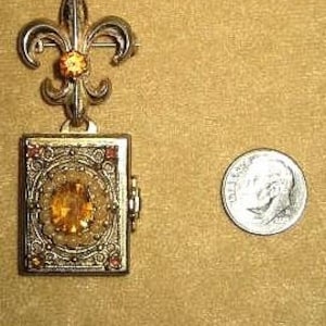 May include: A vintage gold-toned metal brooch with a fleur-de-lis design and a rectangular locket. The locket features an orange gemstone and is next to a U.S. dime for size comparison. The brooch has an ornate, detailed design.