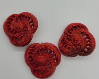 May include: Three red plastic buttons with a decorative, intricate, floral design. The buttons are shaped like a heart with a swirl pattern.