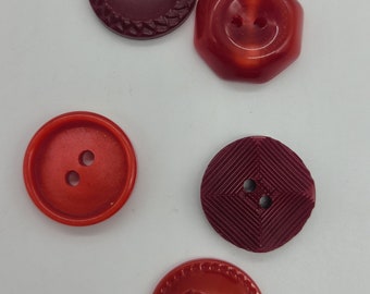 May include: Five plastic buttons in red and burgundy colors. Two buttons have a raised design, two have a smooth surface, and one has a diamond pattern.