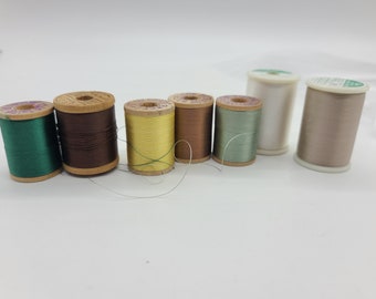 May include: A collection of eight spools of thread in various colors, including green, brown, yellow, brown, green, white, and beige. The spools are made of wood and have a paper label.