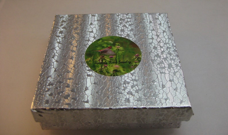 May include: A silver gift box with a round sticker featuring a bird in flight and the text "Sparkle Finch Jewelry".