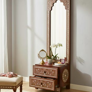 May include: A dark brown vanity unit with a tall, arched mirror. The mirror and drawers feature detailed white inlay patterns. A small round mirror, a tray, and a potted plant sit on top. A matching stool is in the foreground.